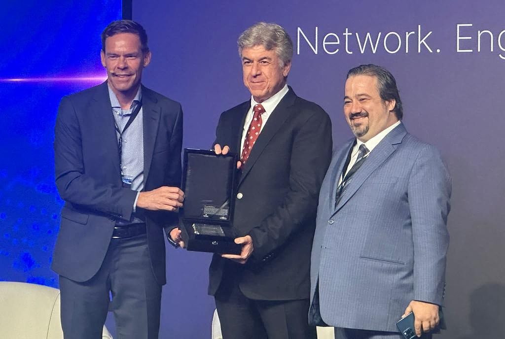 Three men are seen on a presentation stage with a trophy being handed over