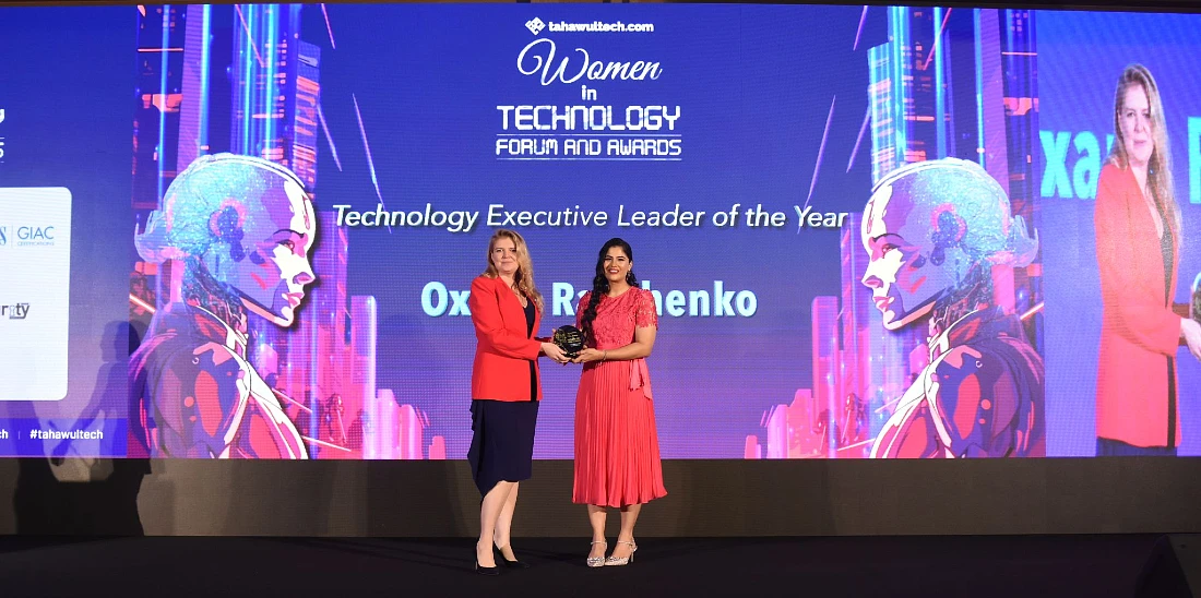 Texte alternatif : une femme vêtue d'une veste rouge tient un trophée devant un écran avec les mots Women in Technology Forum et des récompenses bien visibles.