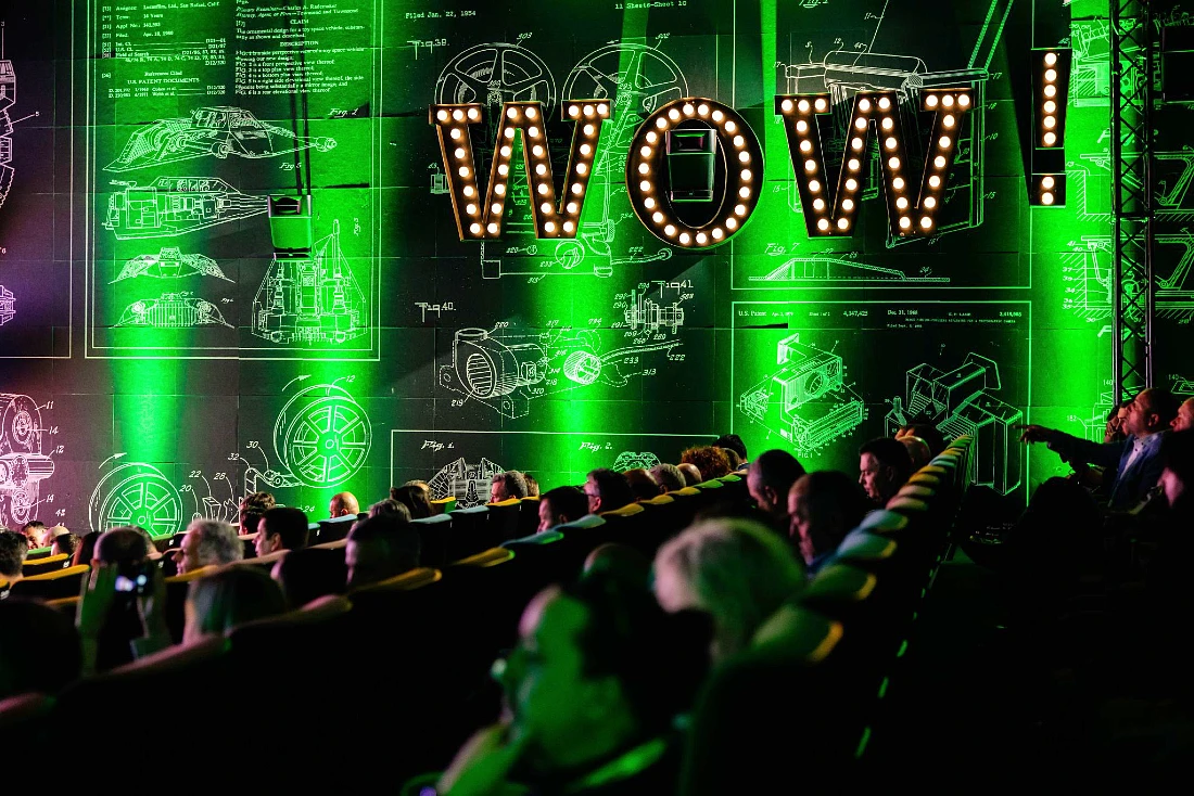A view inside a darkened auditorium filled with people - green uplighters light the dark space and the word Wow! is seen picked out in lights on the side of the space