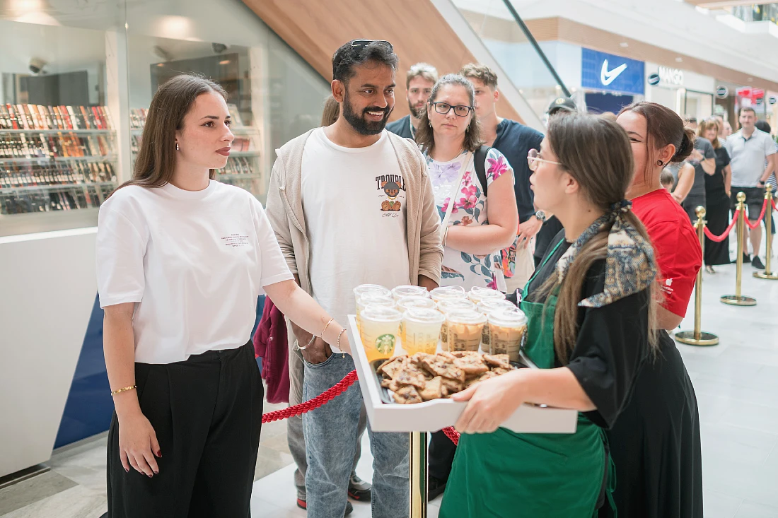 a woman holding a tray of drinks and snacks speaks to two people in a queue in a shopping mall