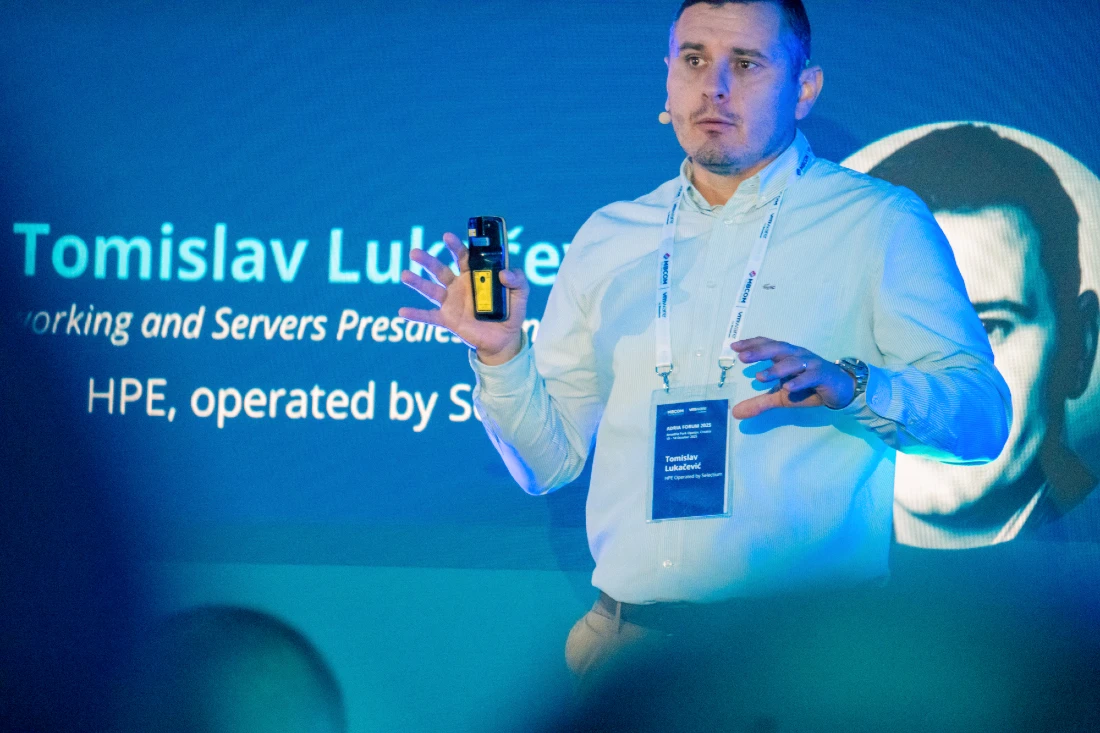 a man in a shirt presents to an audience at a hi tech event, holding a mouse device used to change presentation slides