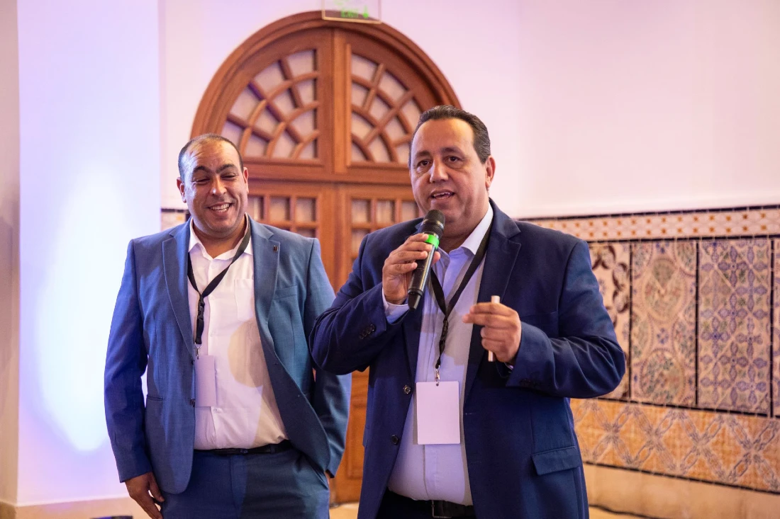 Two men are presenting at a hi tech event. The man in front is speaking into a mic.