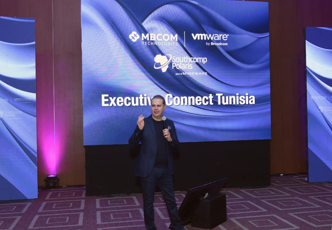 a man is talking into a mic at a hi tech event with a digital projection screen behind him