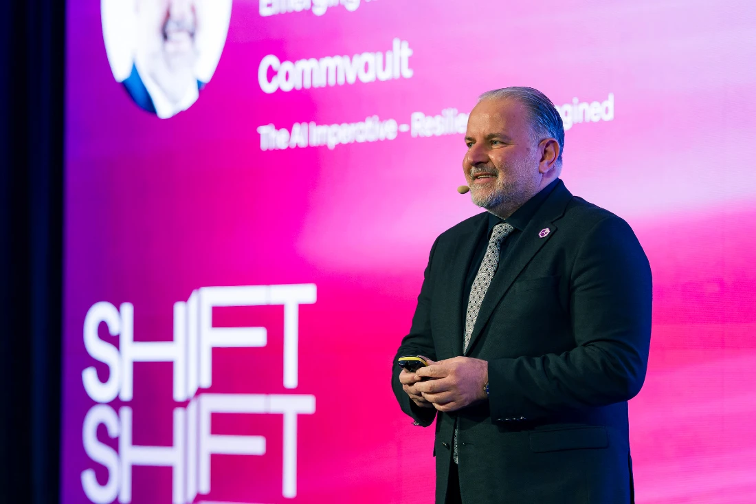 Un homme prend la parole lors d'une conférence high-tech avec un fond rose projeté.