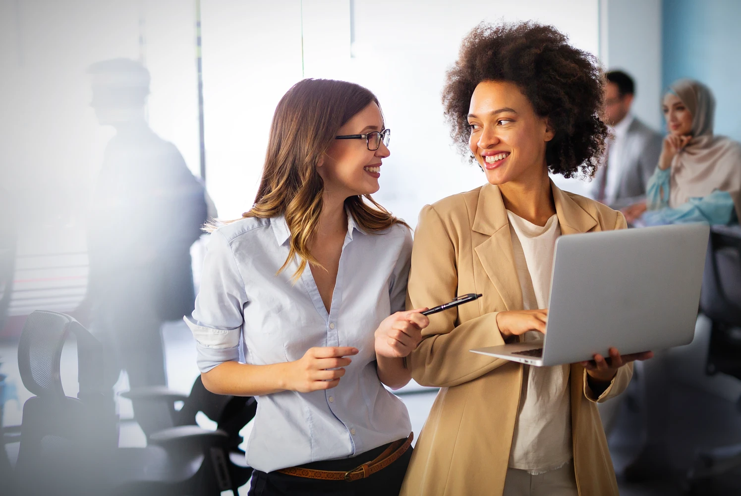 two women are talking to each other and smiling in a office setting. One of them holds a laptop.