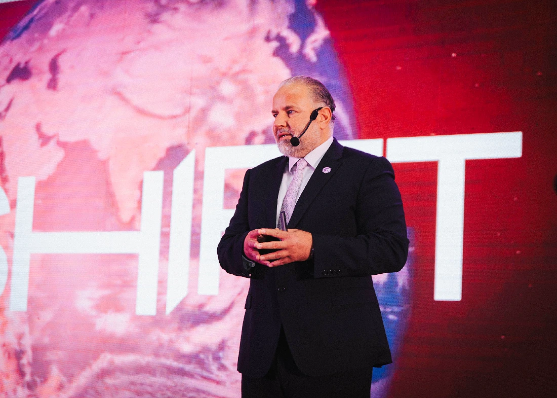 A man in a suit presents to an audience in front of a screen with the word SHIFT visible