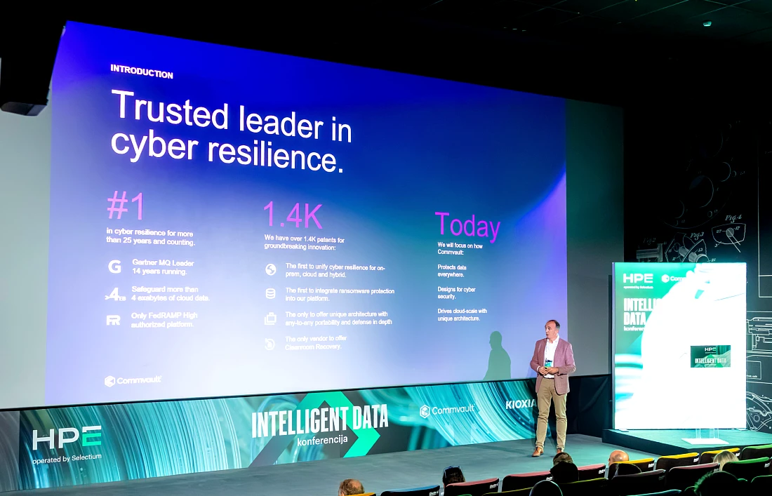 A man presents to an audience at a hi tech event. He looks at a very large presentation screen as he talks