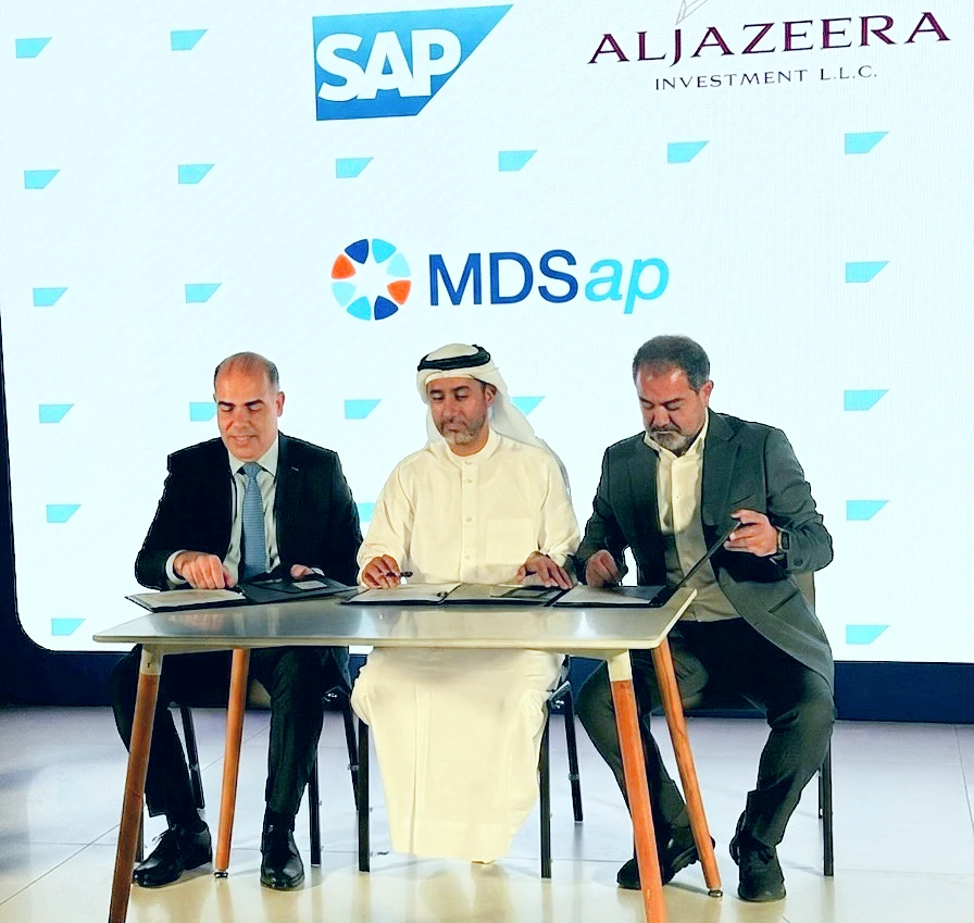 Three men are seen signing a partnership agreement. Behind them, company logos are projected onto a screen.